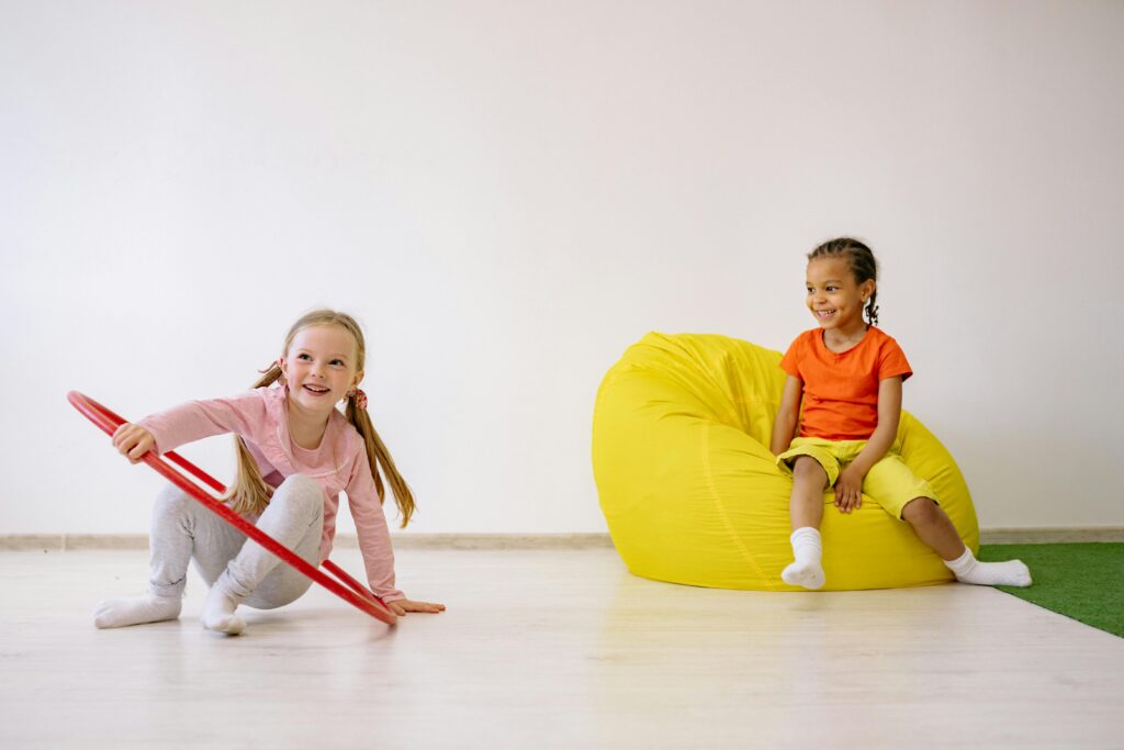 Two children enjoying playtime indoors with a hula hoop and bean bag, full of joy and fun.